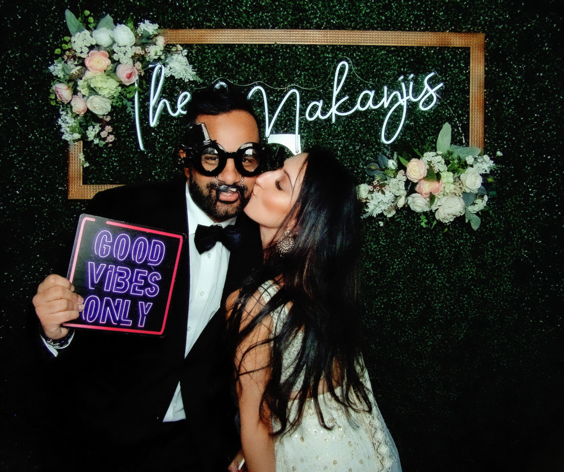 Wedding couple laughing in the photo booth with novelty glasses and a Good Vibes Only sign in front of a greenery wall with a custom neon sign
